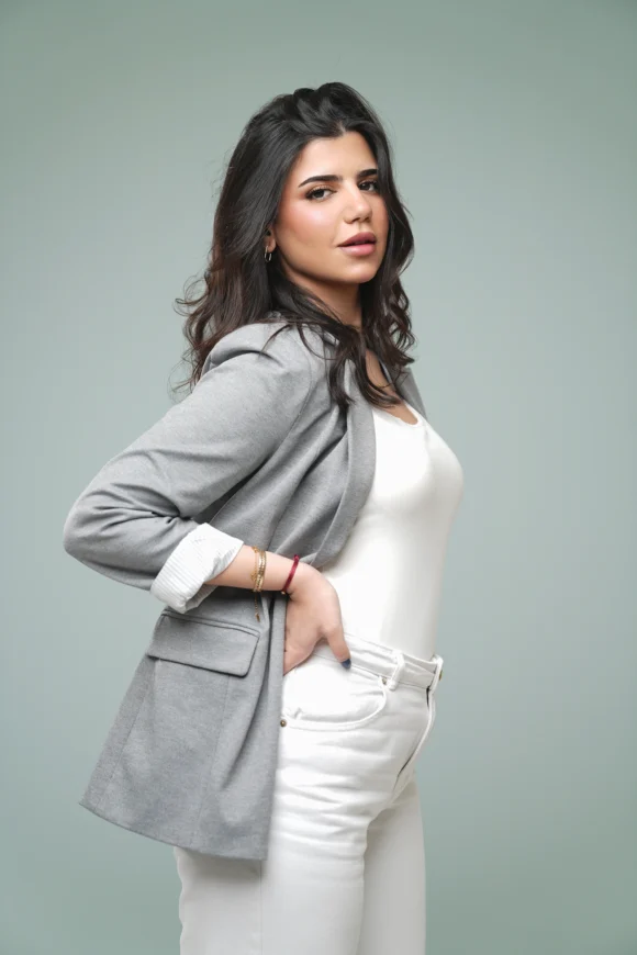 Woman in a gray blazer, white top, and white jeans posing against a light green studio background