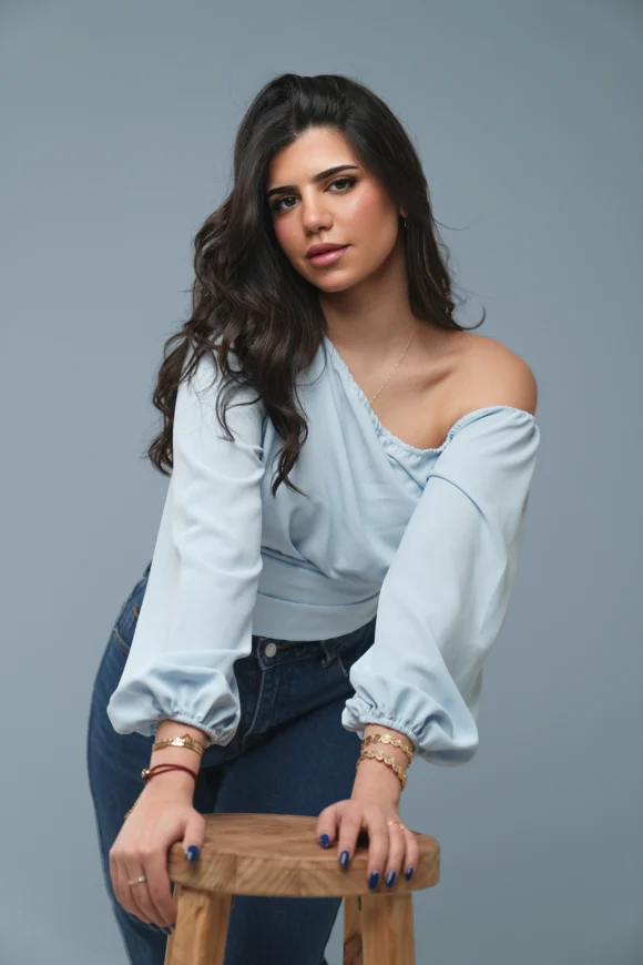 Woman wearing a light blue off-shoulder wrap top, dark jeans, and gold bracelets, leaning on a wooden stool
