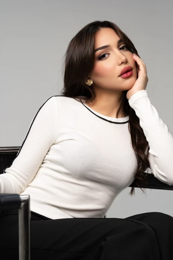 SHD, fashion model in Kuwait with ModelsNova, seated studio portrait in a white top with black trim and black trousers.
