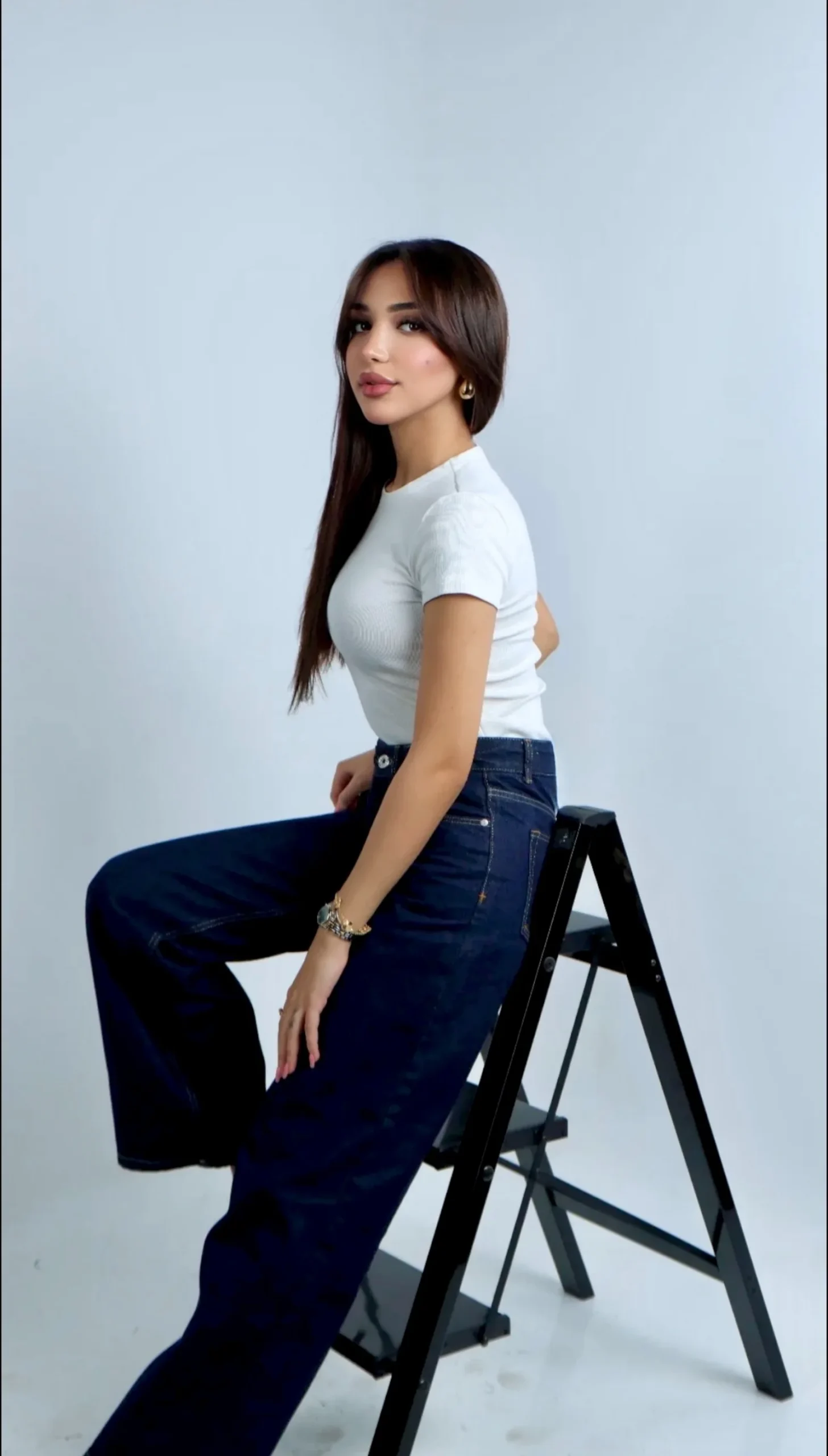 SHD, full length studio shot seated on a step ladder in a white t-shirt and wide leg jeans.