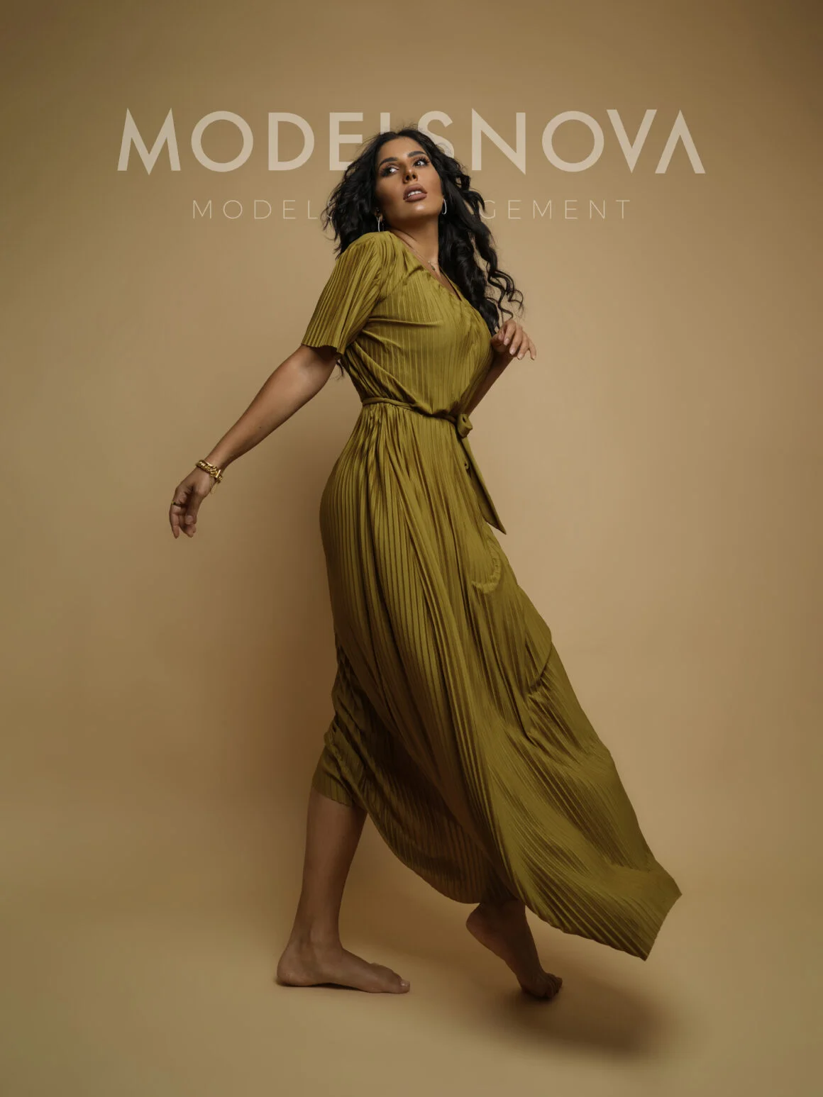 Models Nova promotional photo featuring a woman in a flowing olive pleated maxi dress with gold accessories