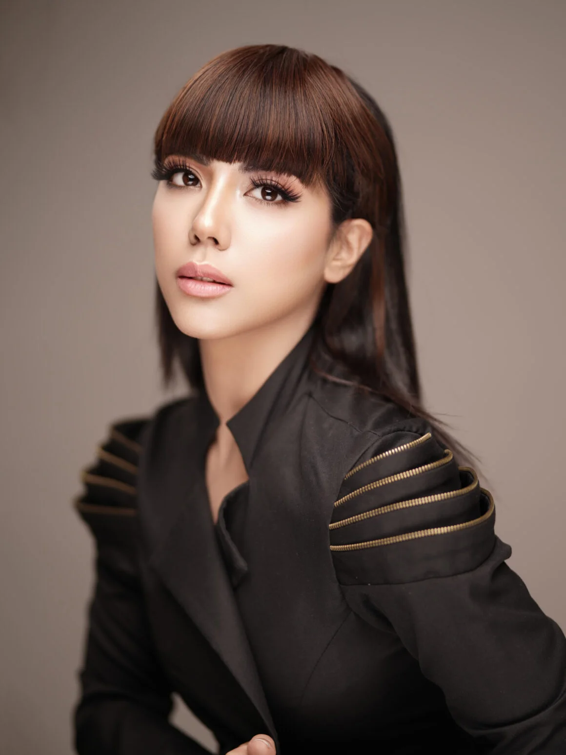 Woman with brown hair and blunt bangs wearing a black jacket with gold zipper shoulder details, studio portrait