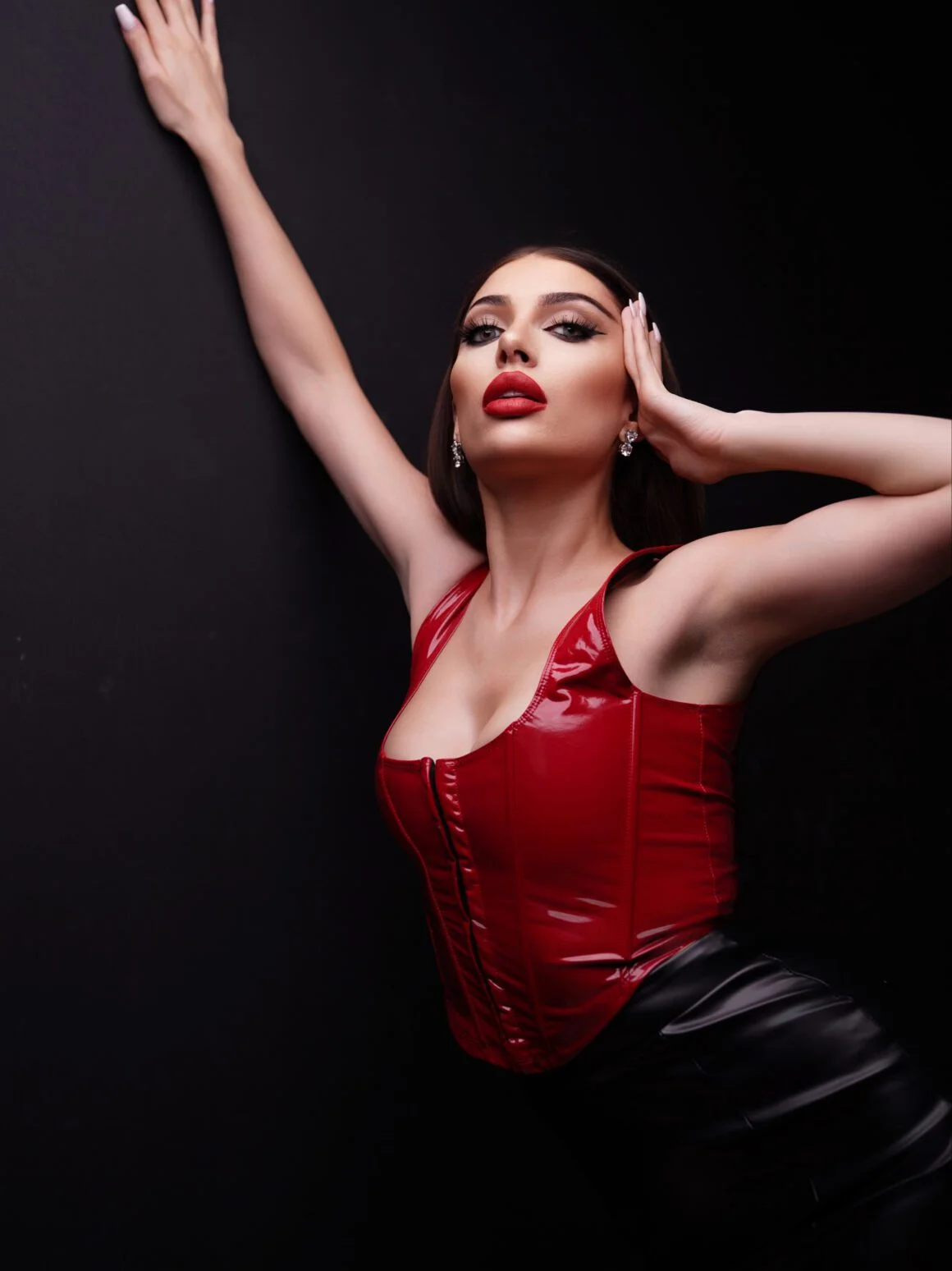 Woman posing in a red patent leather corset and black skirt against a dark background