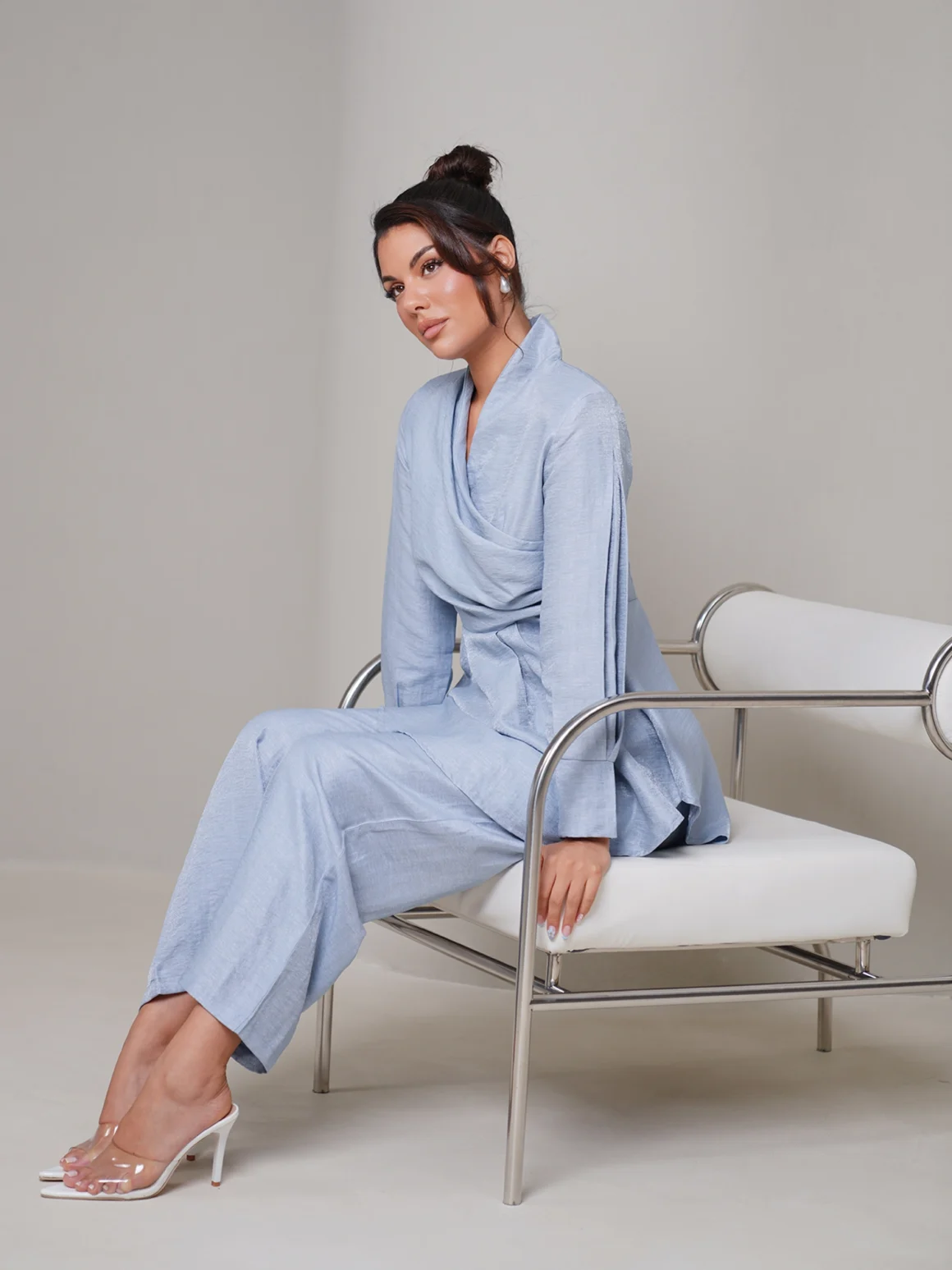 Woman seated on a modern chrome chair wearing a light blue draped linen co-ord set with wide-leg trousers and clear heeled mules