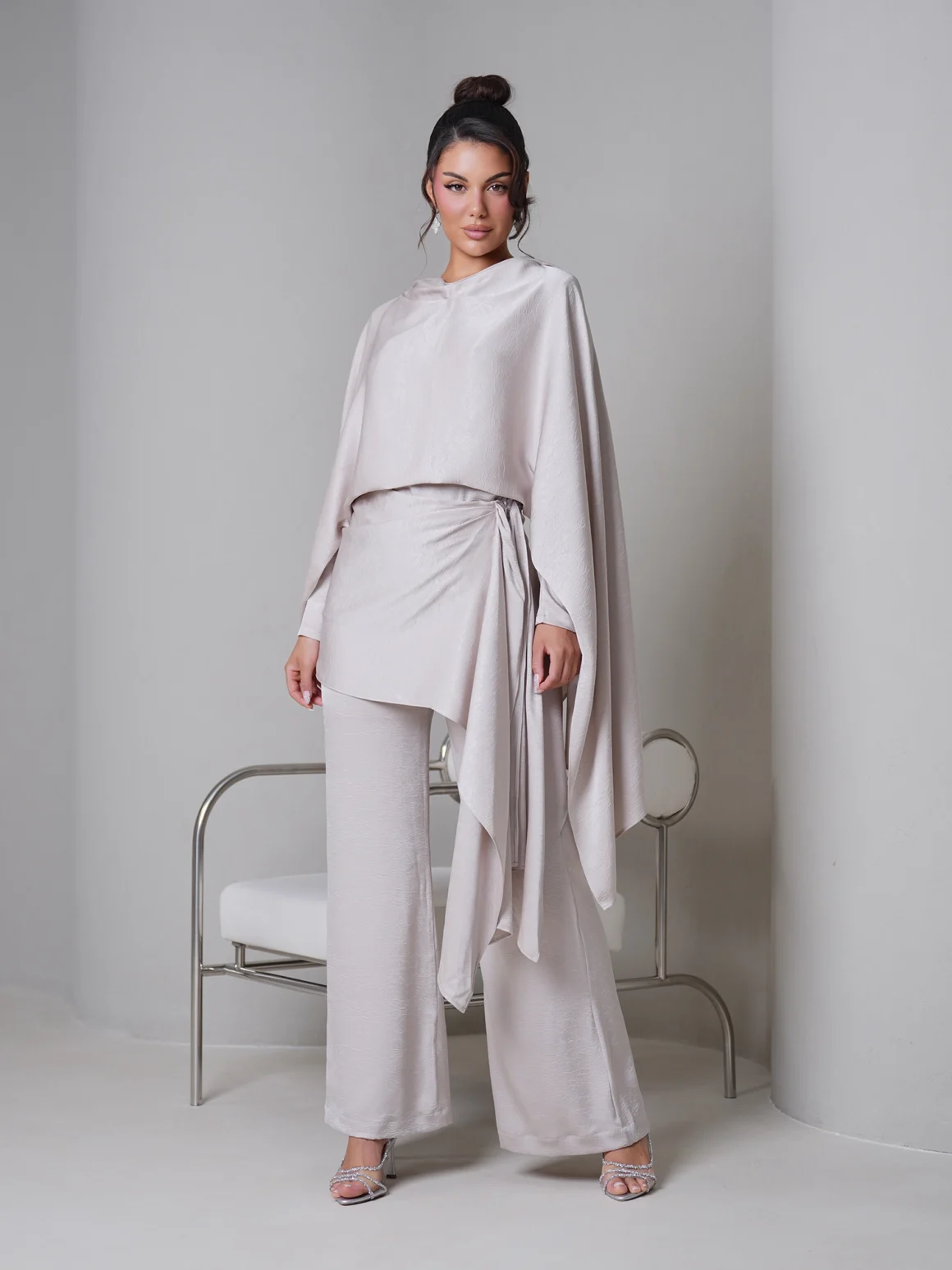 Model wearing a light beige draped cape top with wide-leg pants and strappy heeled sandals