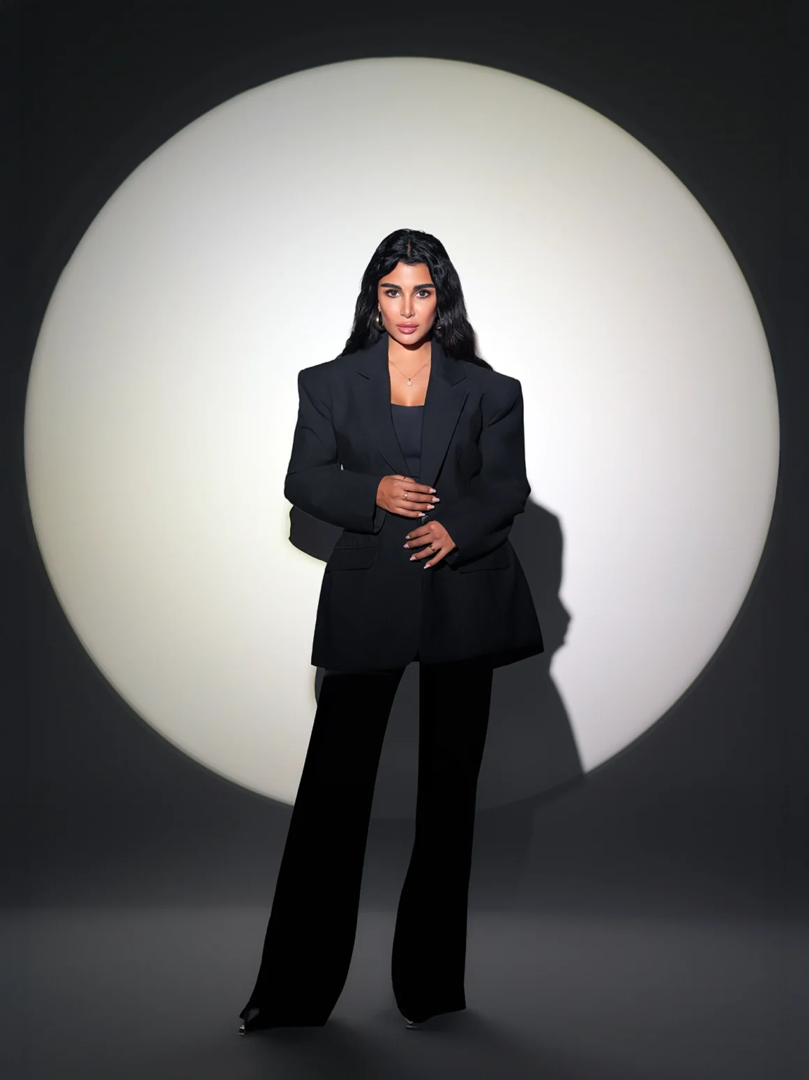 Woman in an oversized black blazer and wide-leg trousers, standing before a large circular light backdrop