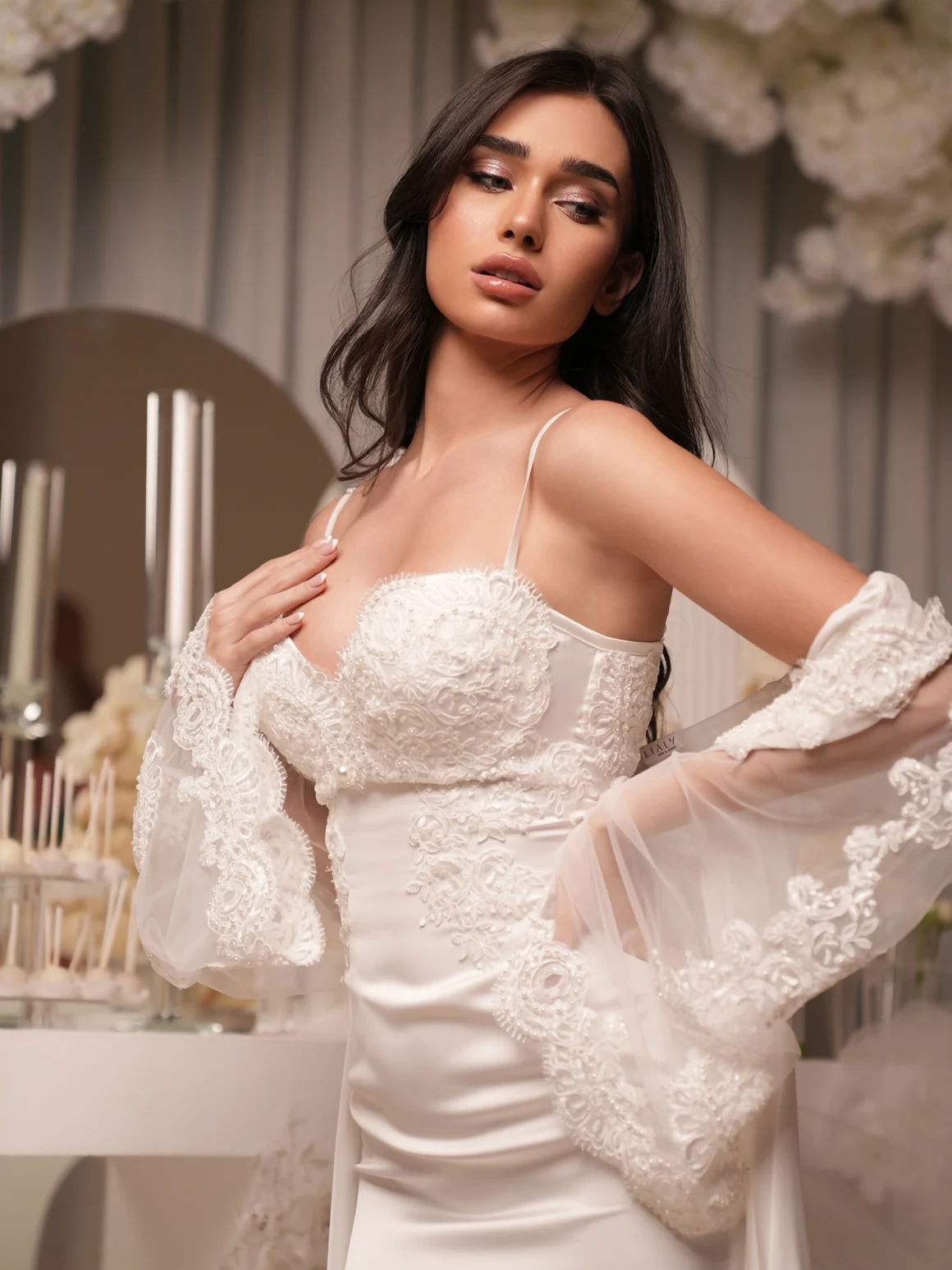 Model wearing a white satin wedding gown with lace appliqué bodice and sheer lace sleeves in a floral-decorated venue