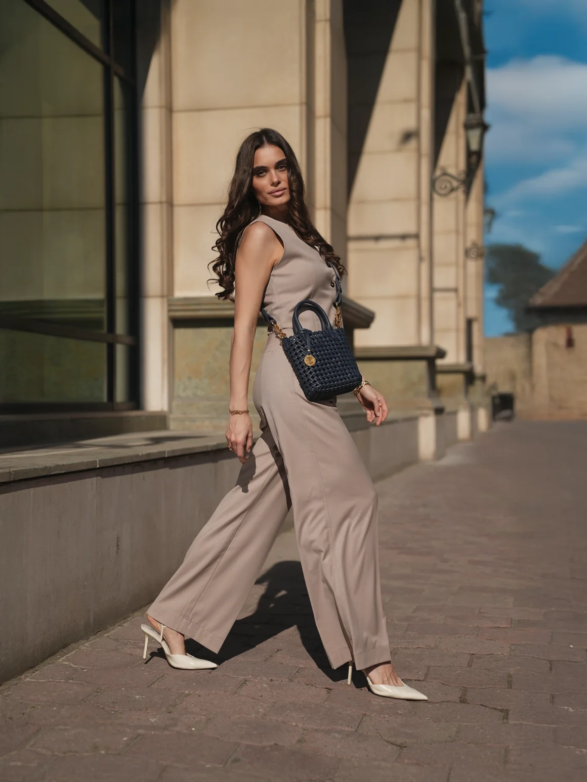 Woman in a beige sleeveless jumpsuit and white heels carrying a small navy woven bucket bag with gold hardware outdoors